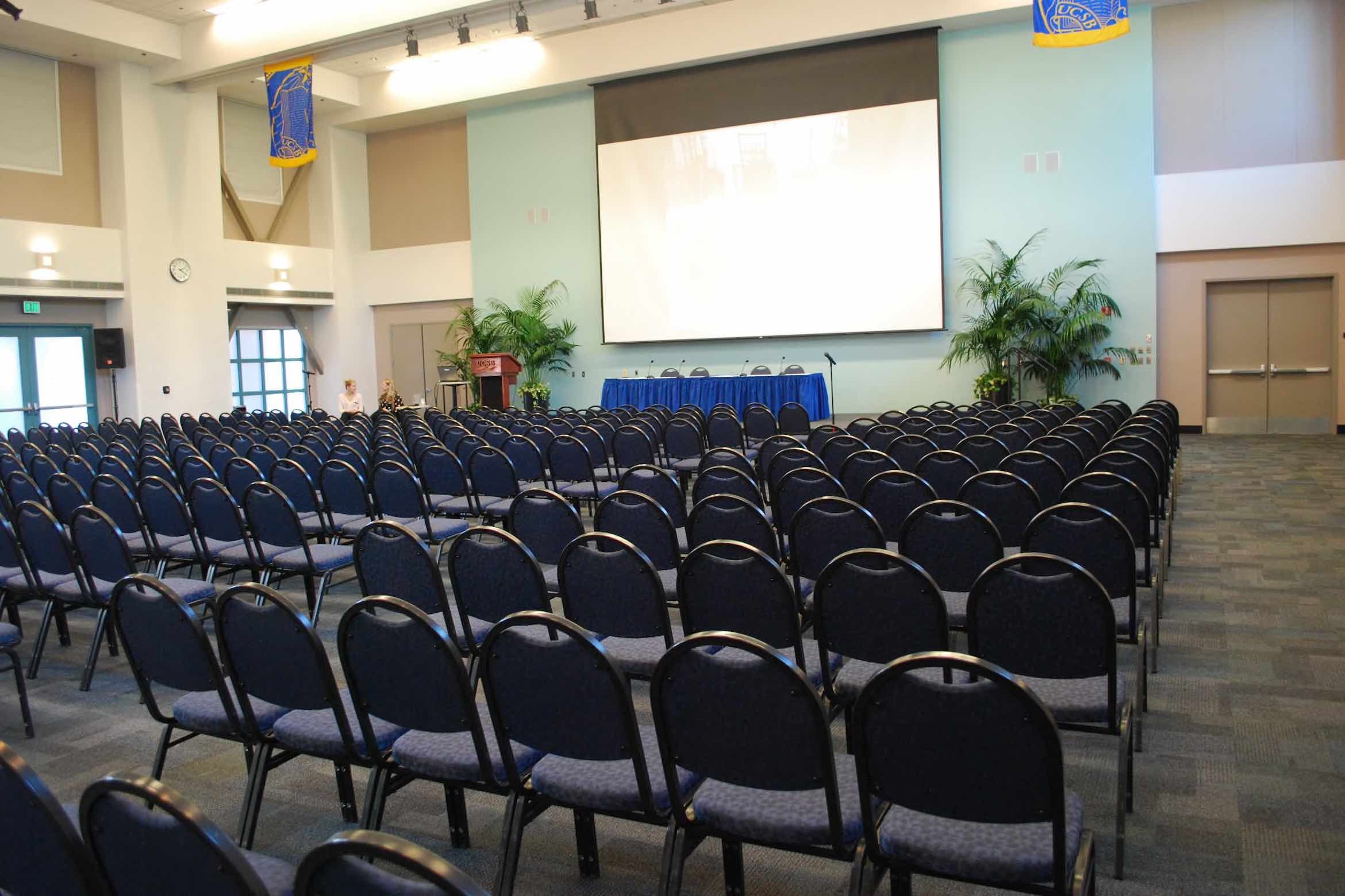 An empty conference room with rows of chairs facing a stage, a podium, and a large screen, ready for a presentation or event.
