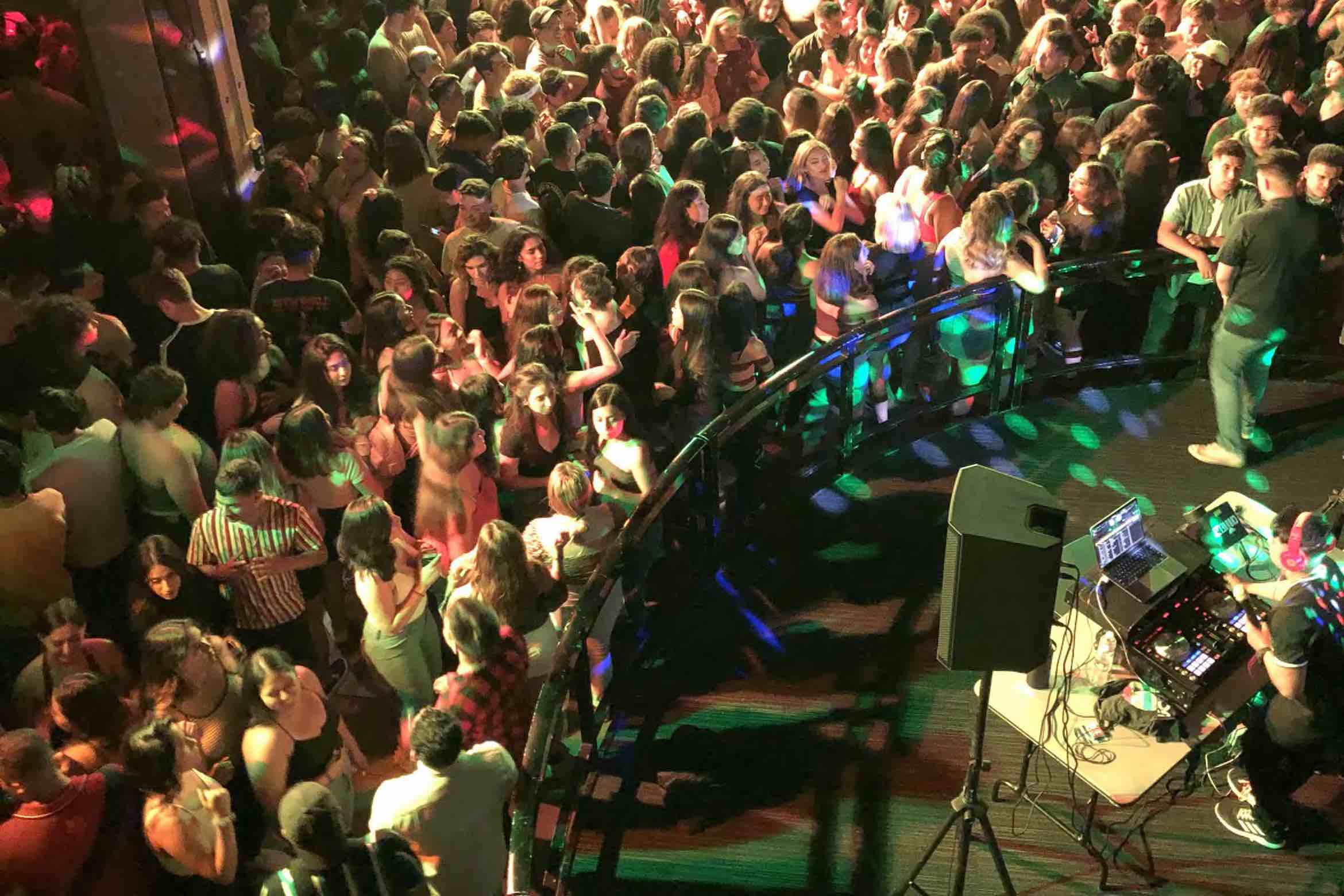 A lively nightclub scene with a DJ playing music as a large crowd dances under colorful lights, creating an energetic atmosphere.