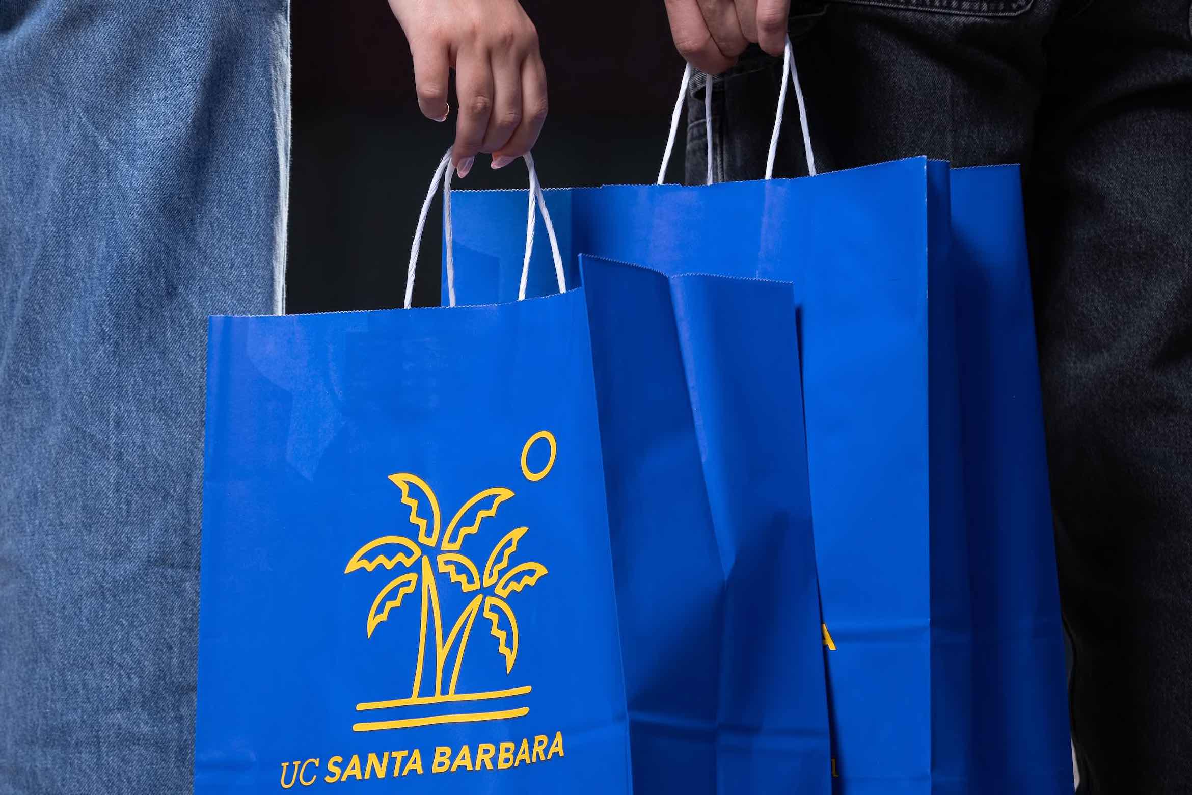 Two people hold bright blue UC Santa Barbara shopping bags with yellow palm tree logos, standing side by side in casual jeans.