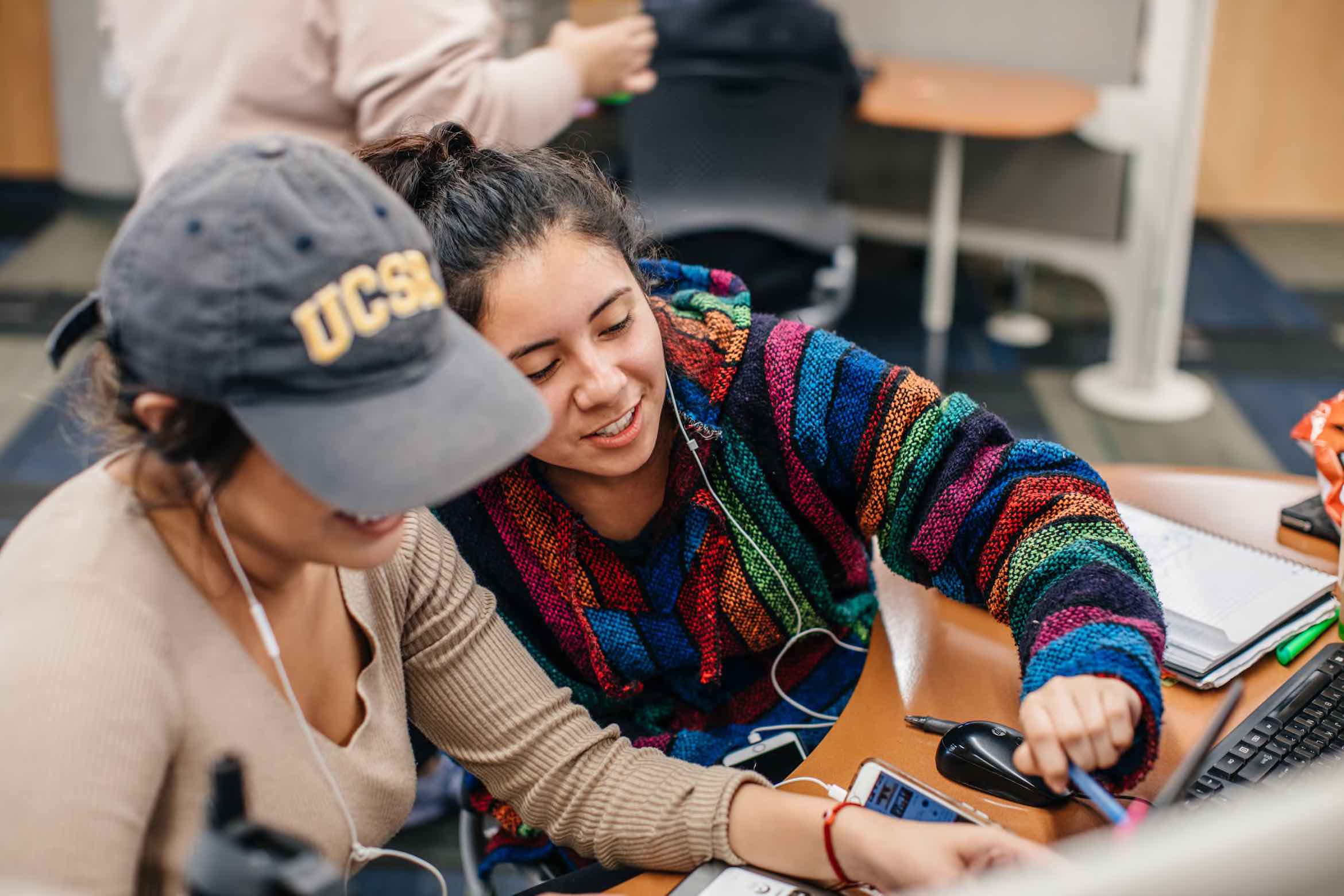 Two students studying together, one in a UCSB cap and the other in a colorful hoodie, smiling as they work on a laptop in a study space.