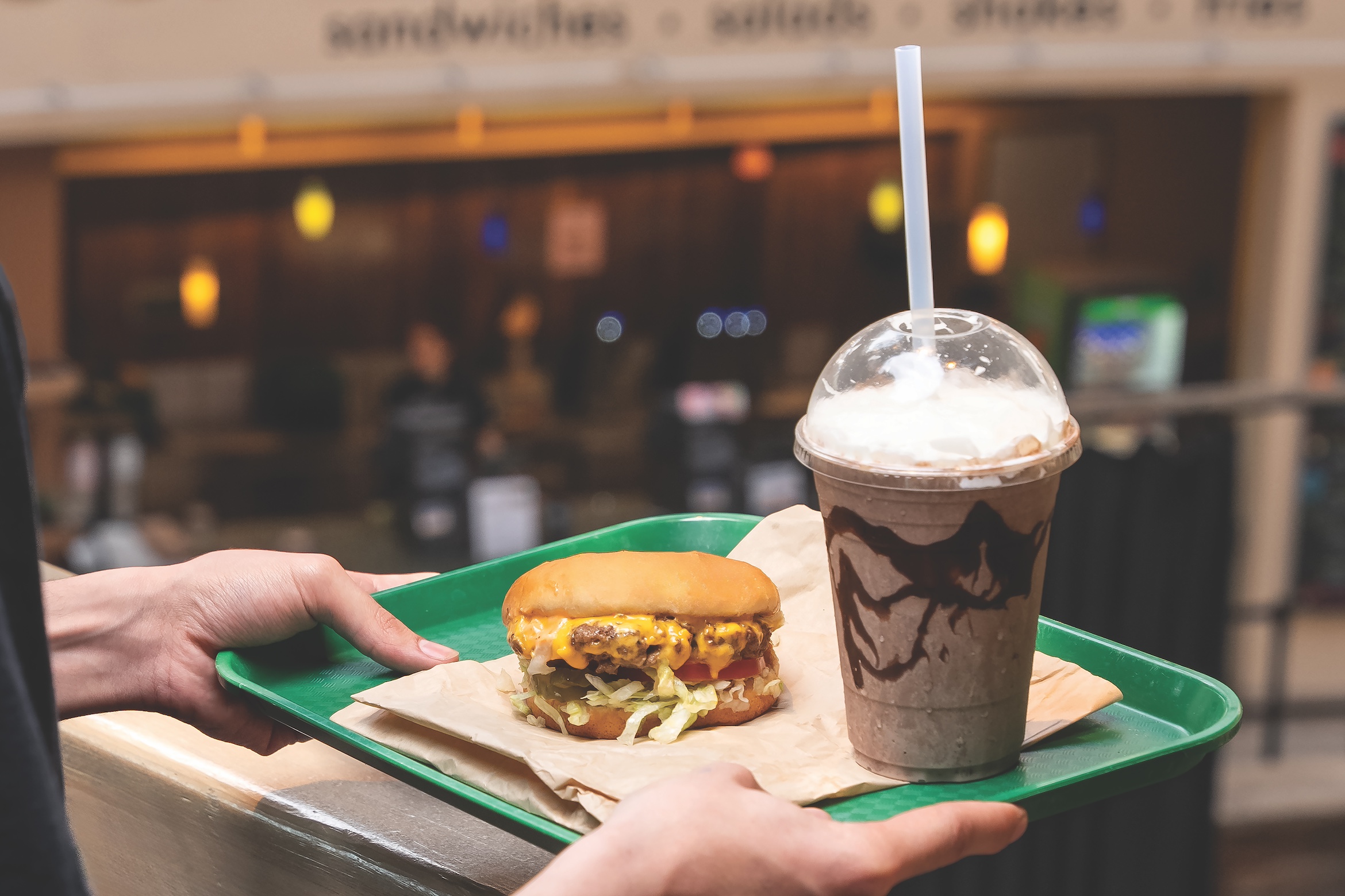 A hand holds a tray with a cheeseburger loaded with lettuce and a whipped cream-topped chocolate milkshake with syrup drizzle.