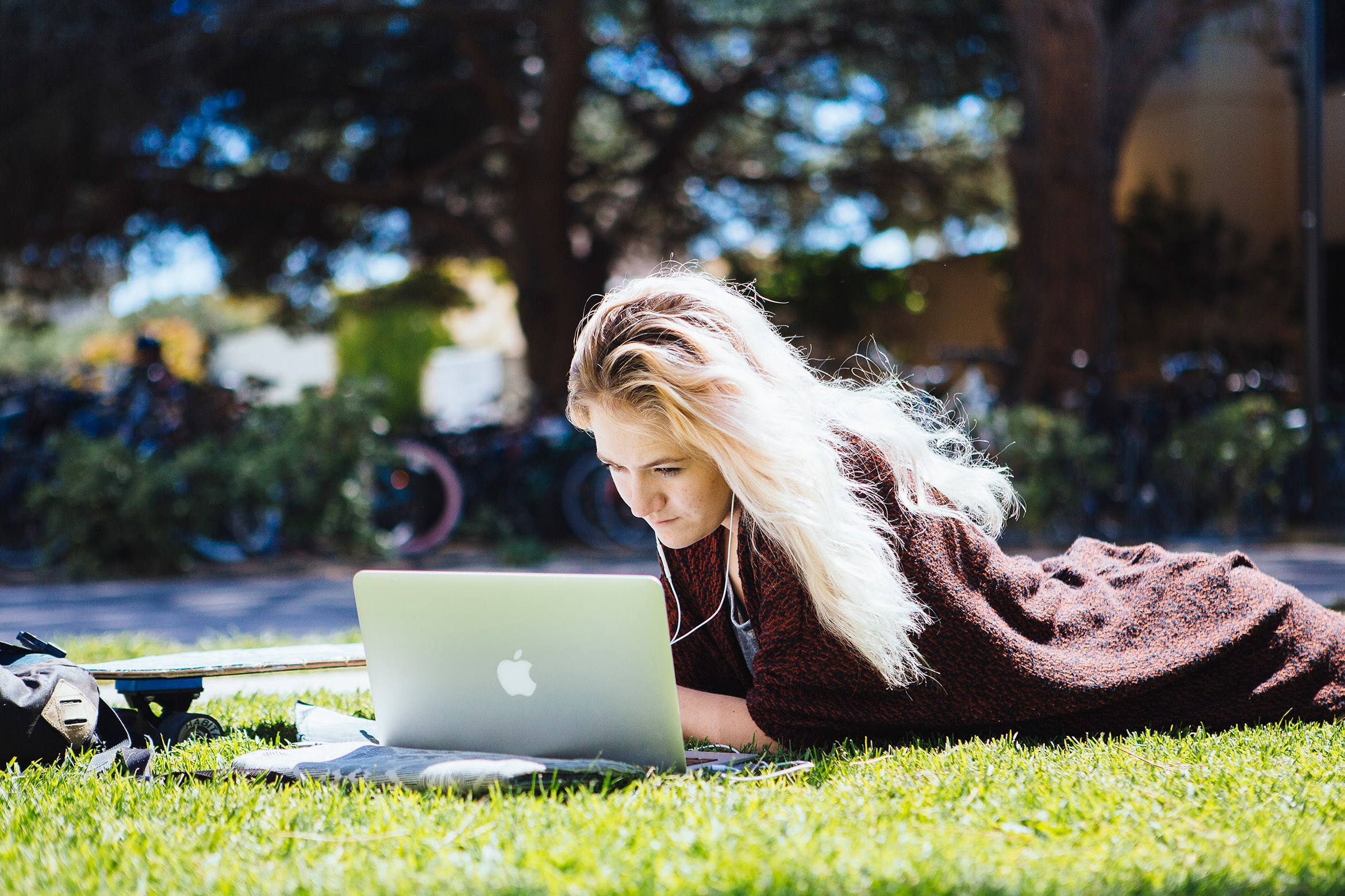 A student lies on the grass with a laptop, focused and studying under dappled sunlight, earbuds in, skateboard and bag nearby.