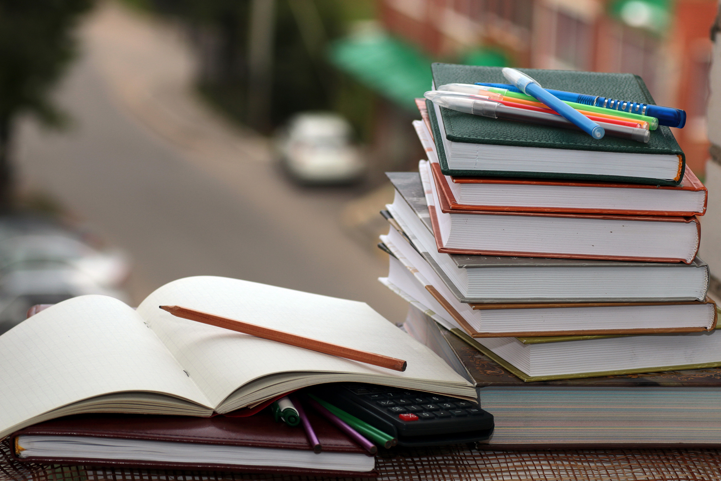A stack of books, pens, and an open notebook with a pencil and calculator rest on a table overlooking a blurred street scene.