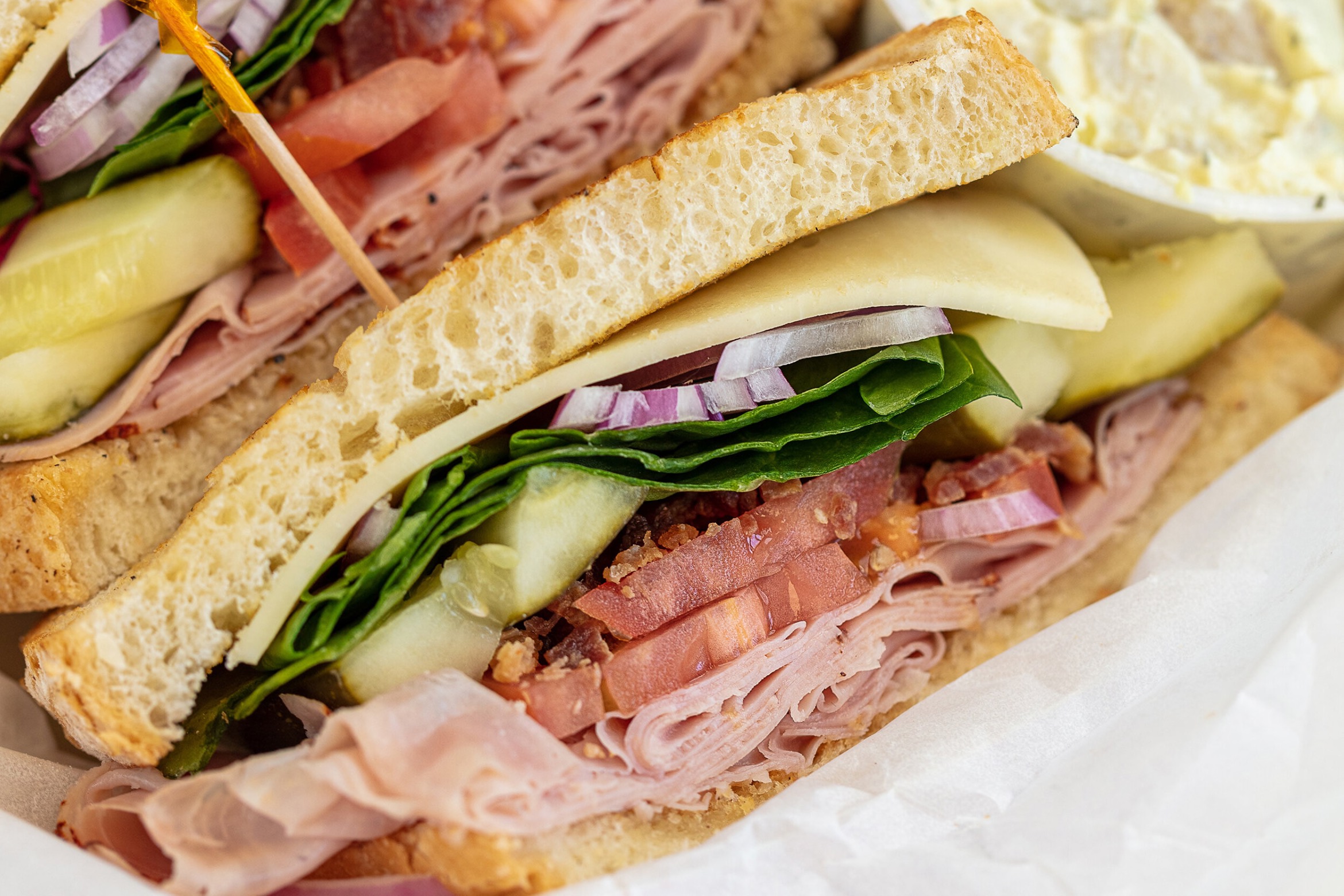 A deli sandwich packed with ham, cheese, tomato, pickles, onions, and greens on thick bread, served with a side of potato salad.
