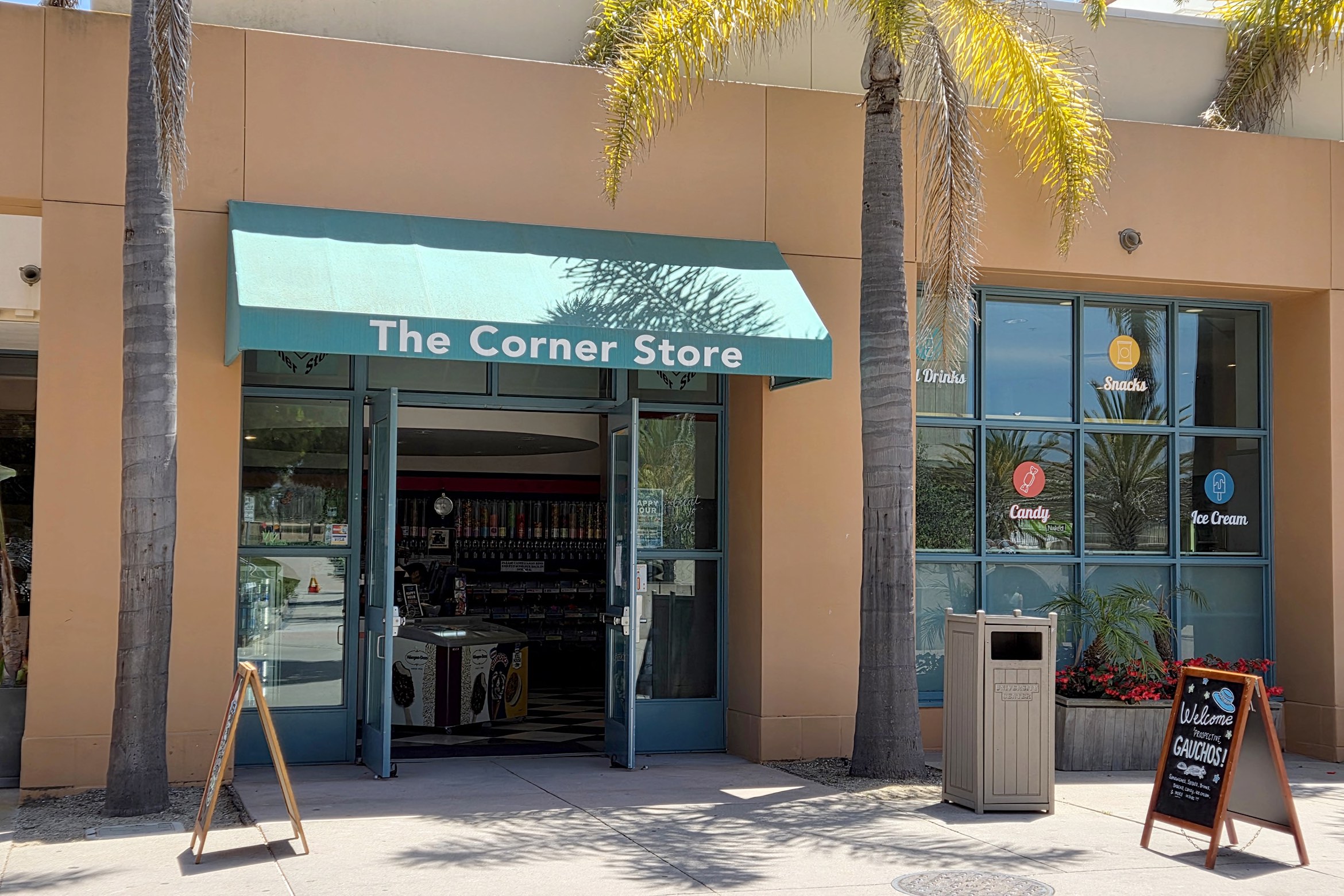 The Corner Store with a teal awning, palm trees outside, and signs for drinks, snacks, candy, and ice cream on the window.