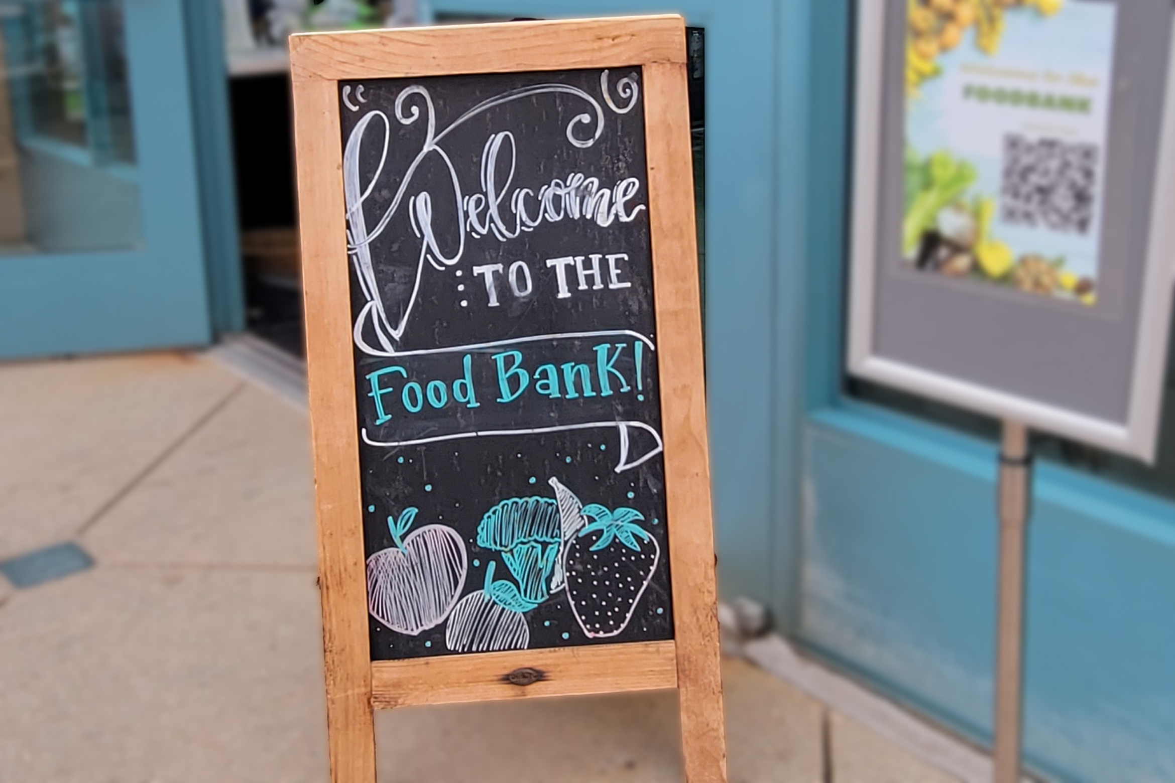 A chalkboard sign with fruit drawings reads "Welcome to the Food Bank!" outside a building with blue doors.