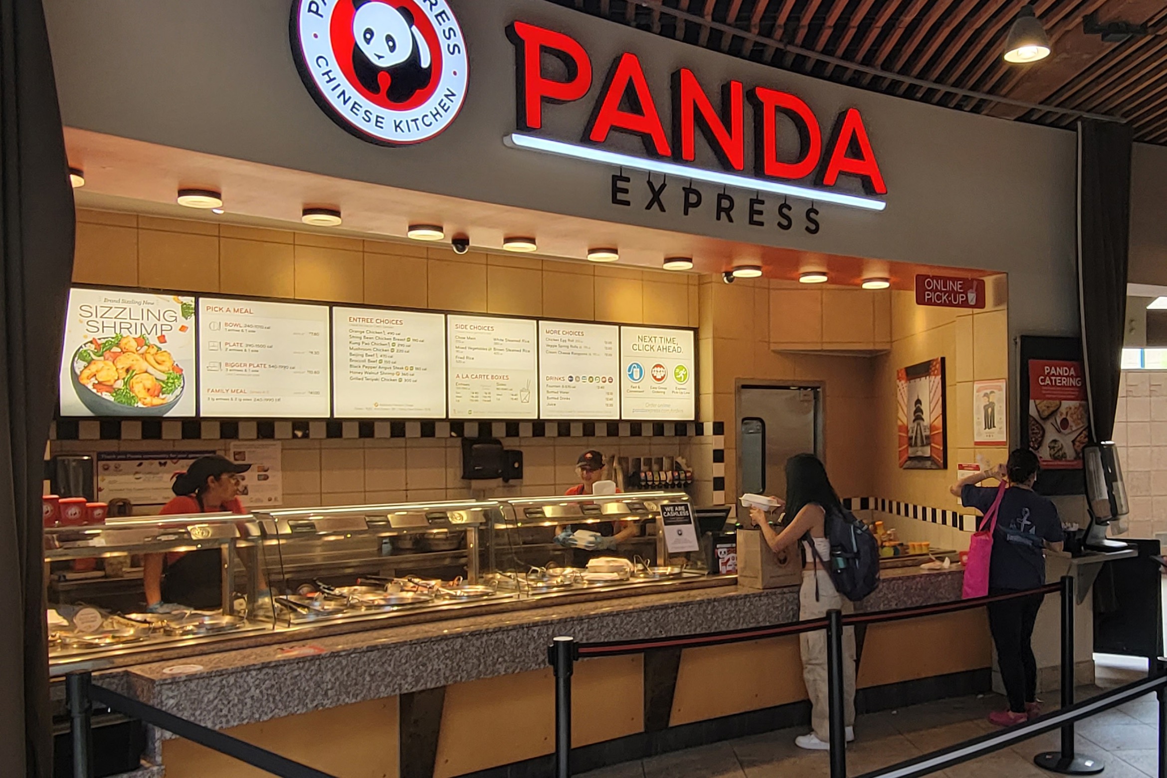 Panda Express counter with staff serving food, two customers waiting, and a lit menu overhead in a mall or food court setting.