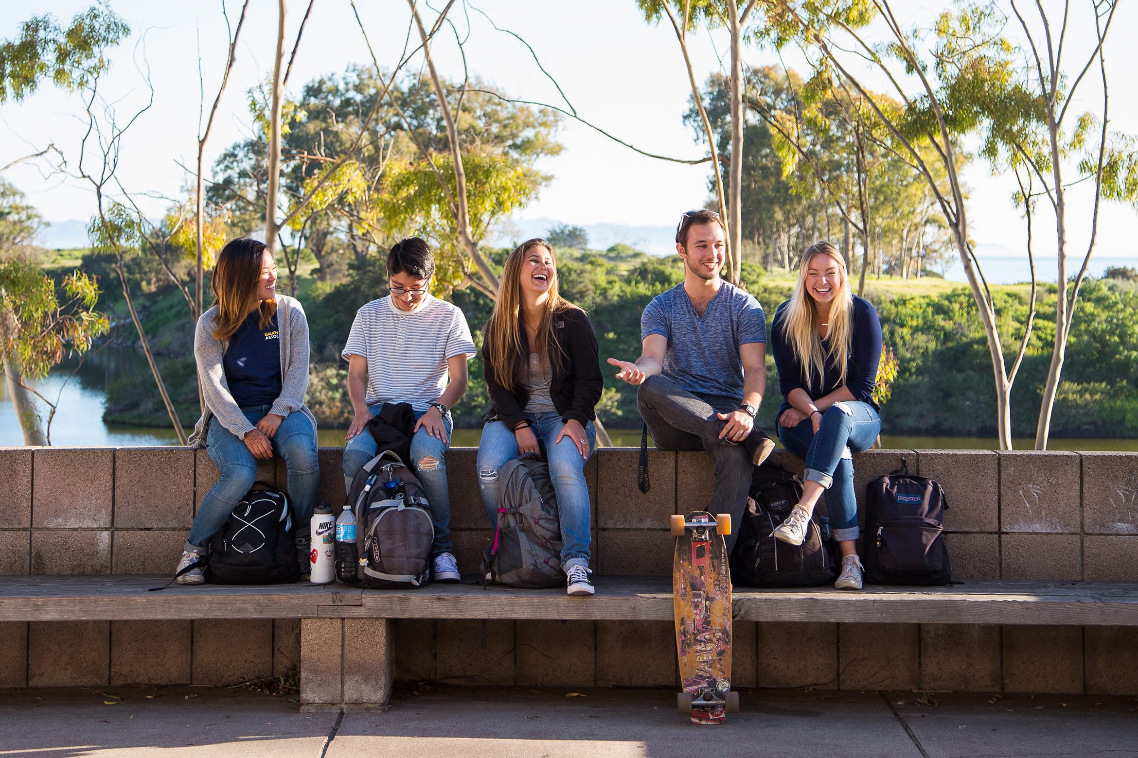 Five college students sit on a bench, laughing and chatting on a sunny day, with backpacks and a skateboard at their feet.