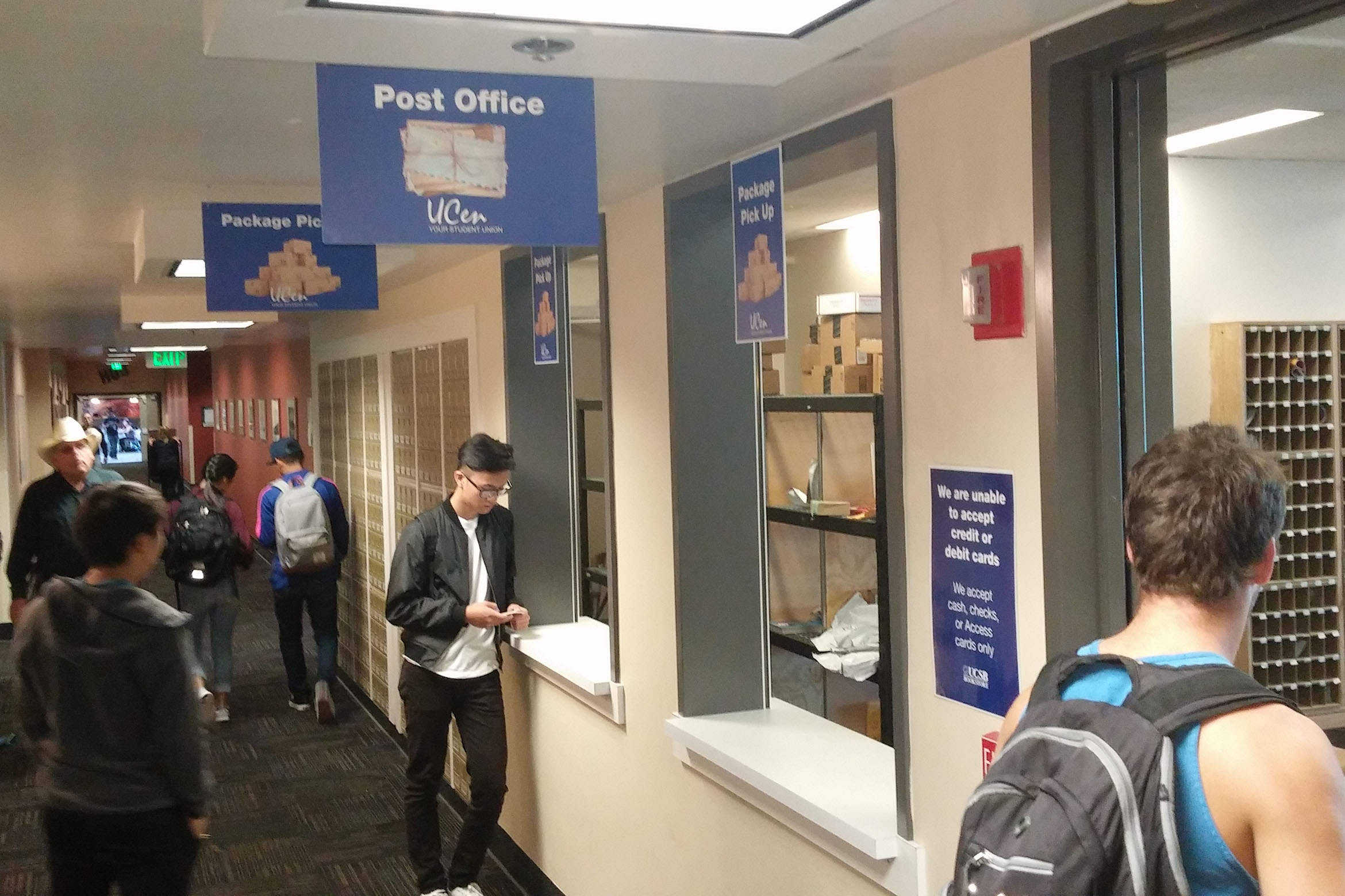Students wait at a campus post office window for package pickup; mailboxes line the hallway in a busy but orderly setting.