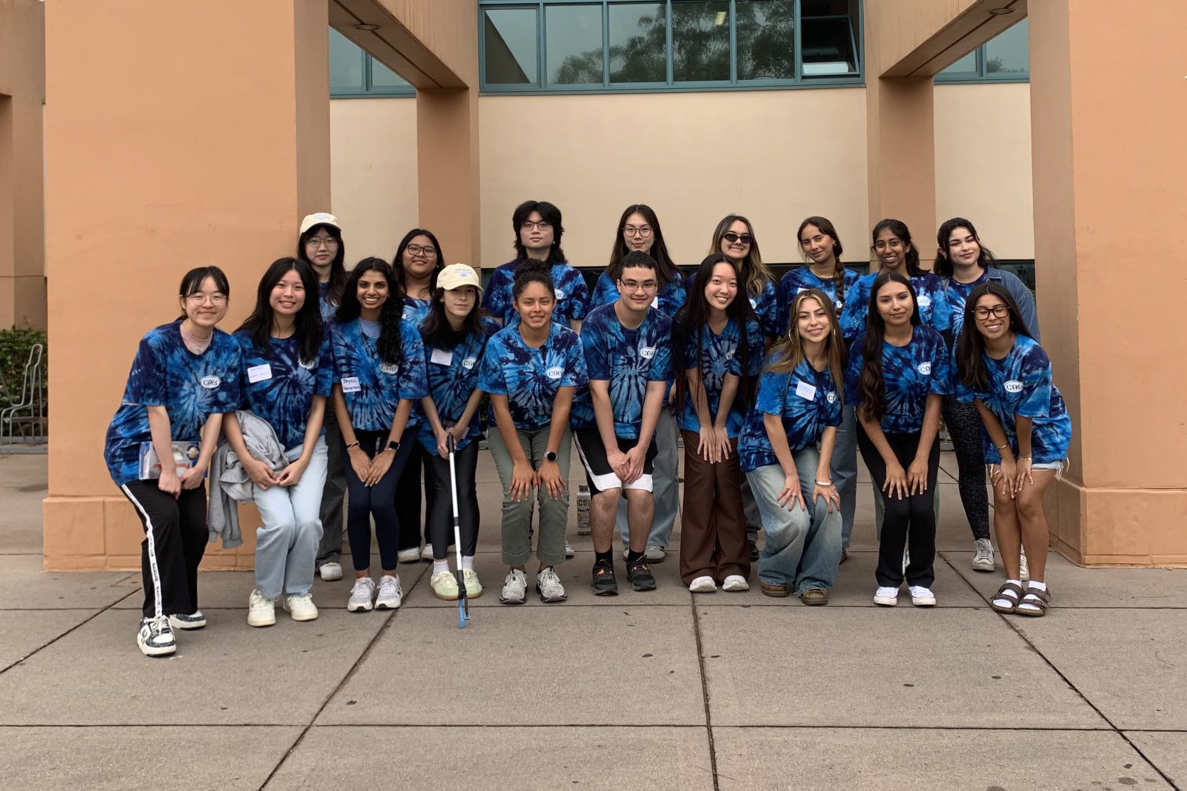 A cheerful group of students in matching blue tie-dye shirts poses outside a campus building, suggesting a club or team event.