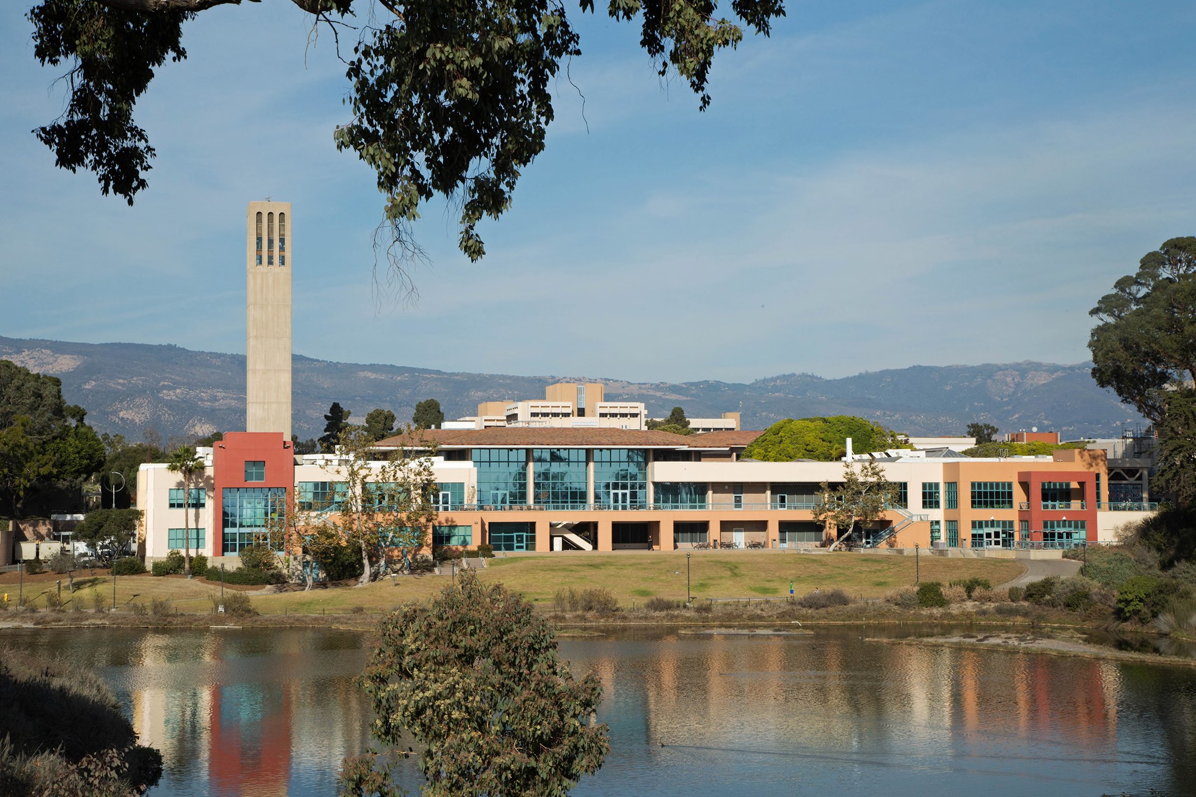 A modern campus building with a tall clock tower stands by a calm lagoon, framed by mountains and blue sky.
