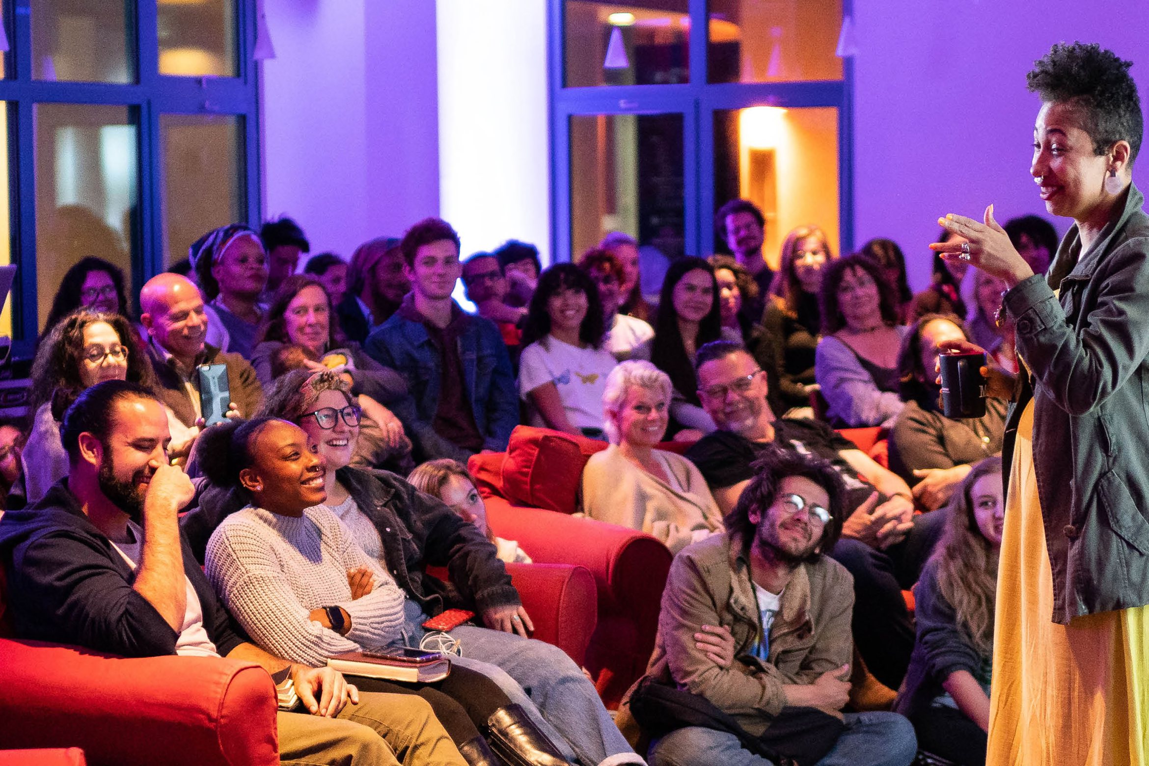 A lively speaker engages a smiling, diverse audience in a cozy, brightly lit room with warm, colorful lighting.
