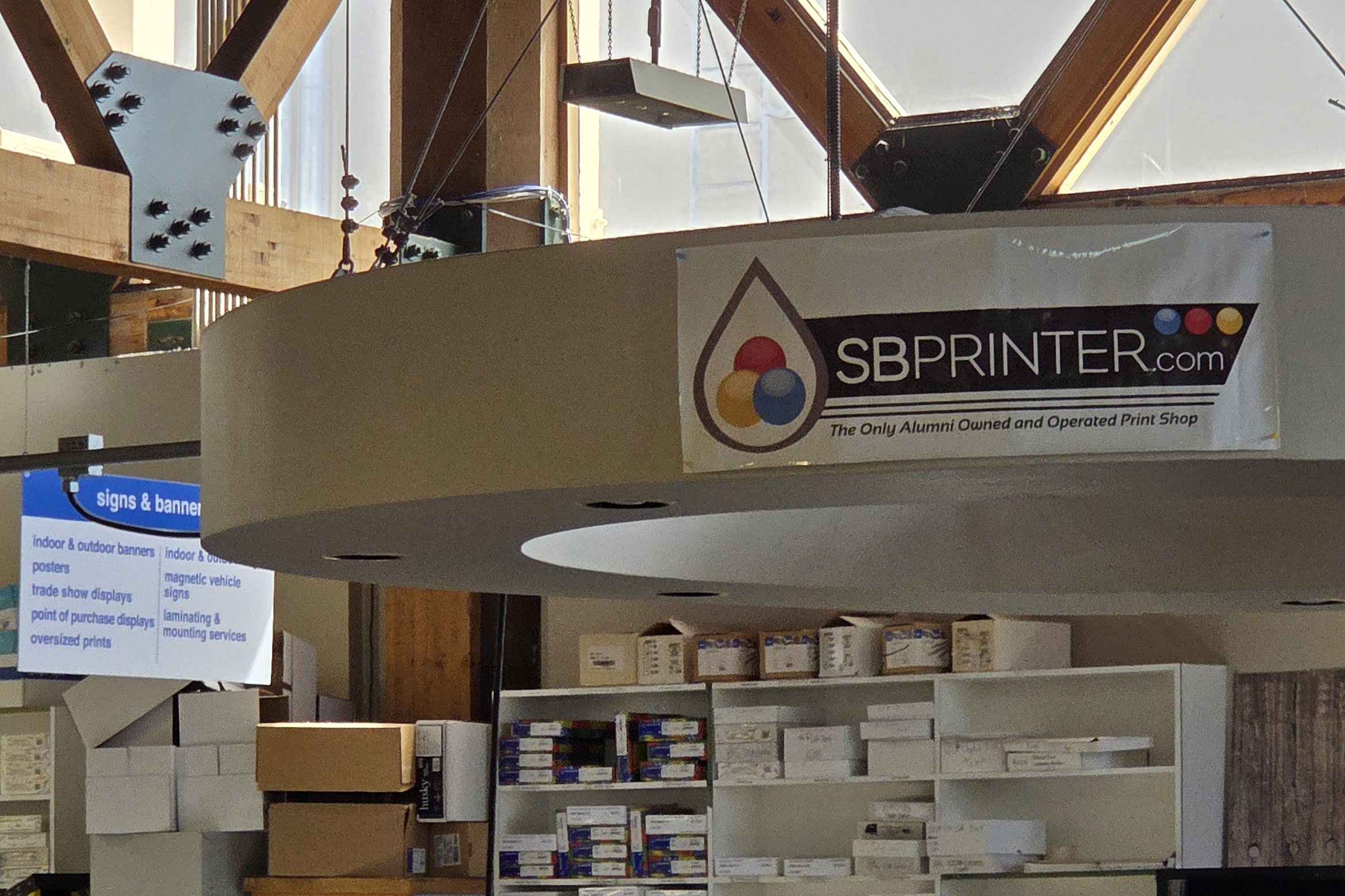 Interior of a print shop with a banner reading “SBPRINTER.com – The Only Alumni Owned and Operated Print Shop.”